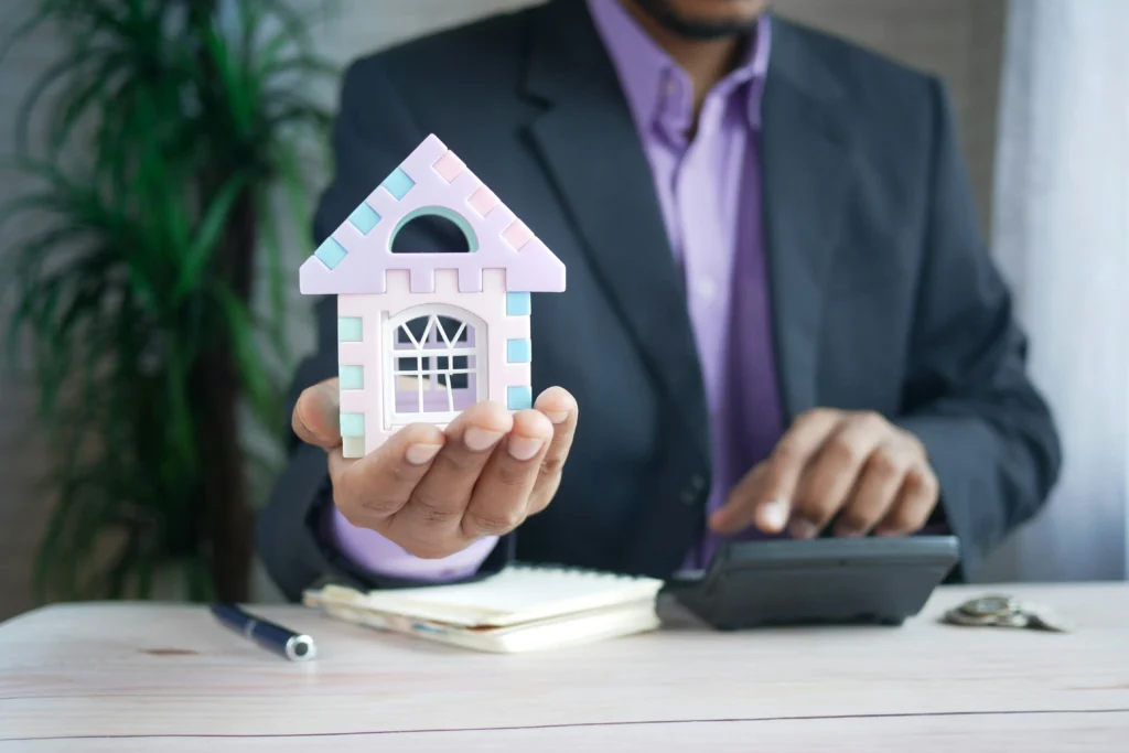 A businessman in a suit holding a small pastel-colored house model while using a calculator, symbolizing real estate investment and financial planning.