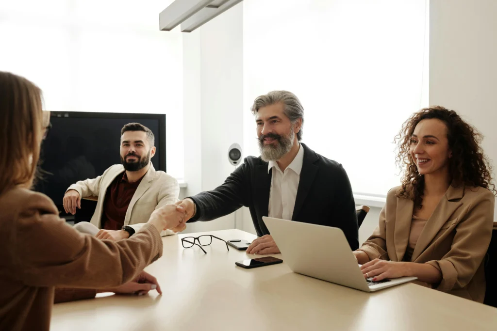 A group of professionals in a modern office, with a smiling businessman shaking hands with a client, symbolizing a successful business deal or partnership.